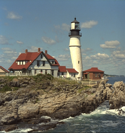 Houses with a lighthouse on top of a cliff above sea, narrower but with a cut in the lighthouse.